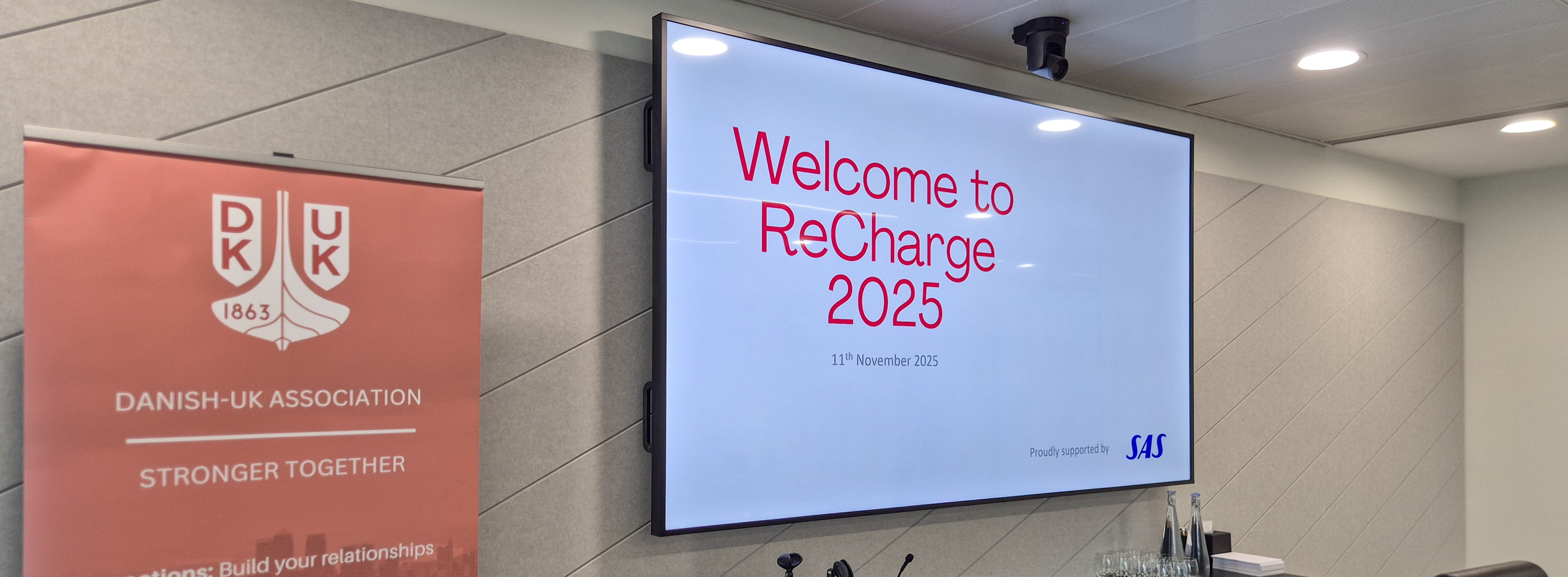 A conference room display showing the welcome slide for “ReCharge 2025” dated 11 November 2025, alongside a Danish‑UK Association banner with the slogan “Stronger Together.” The event is proudly supported by Scandinavian Airlines (SAS).
