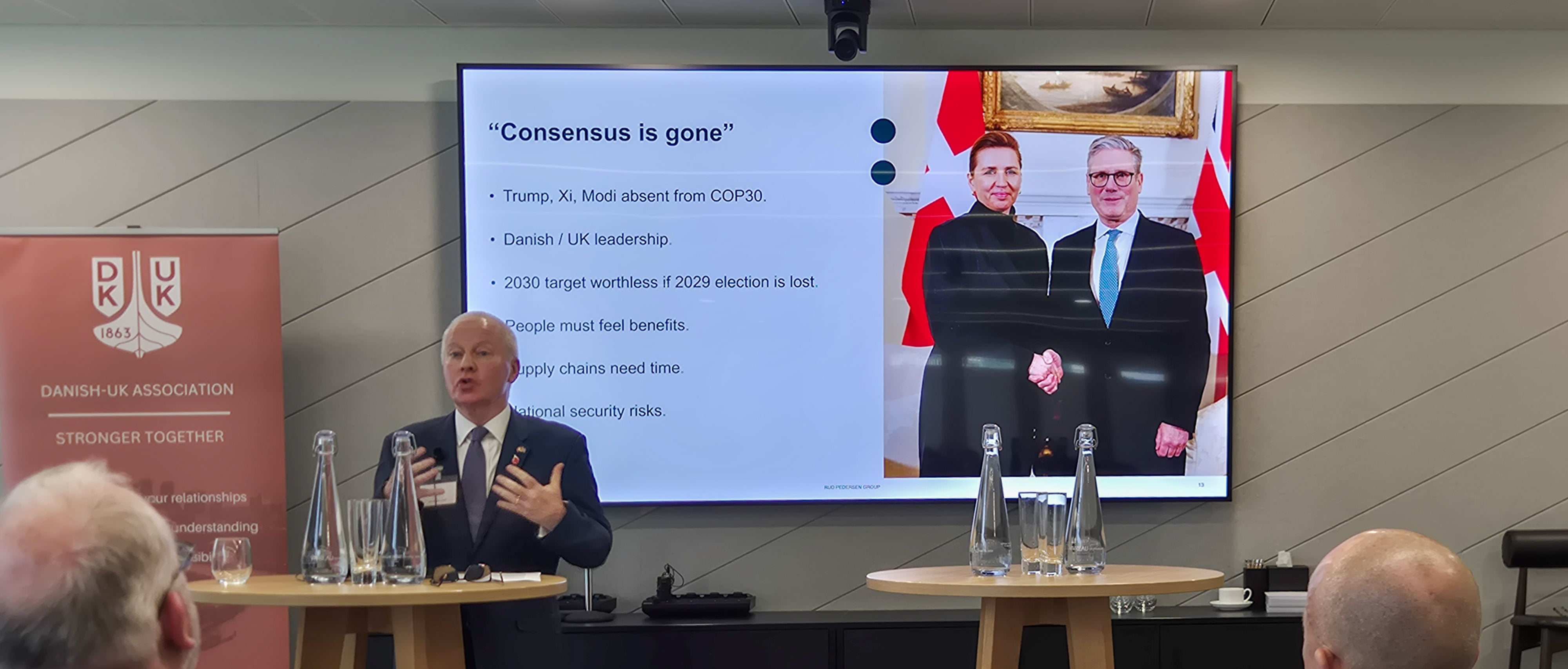 A speaker from the Rud Pedersen Group presents at the ReCharge 2025 event in London, standing beside a Danish‑UK Association banner. On the screen behind him, a slide titled “Consensus is gone” lists geopolitical and policy challenges, alongside a photo of Danish Prime Minister Mette Frederiksen shaking hands with UK Prime Minister Keir Starmer.